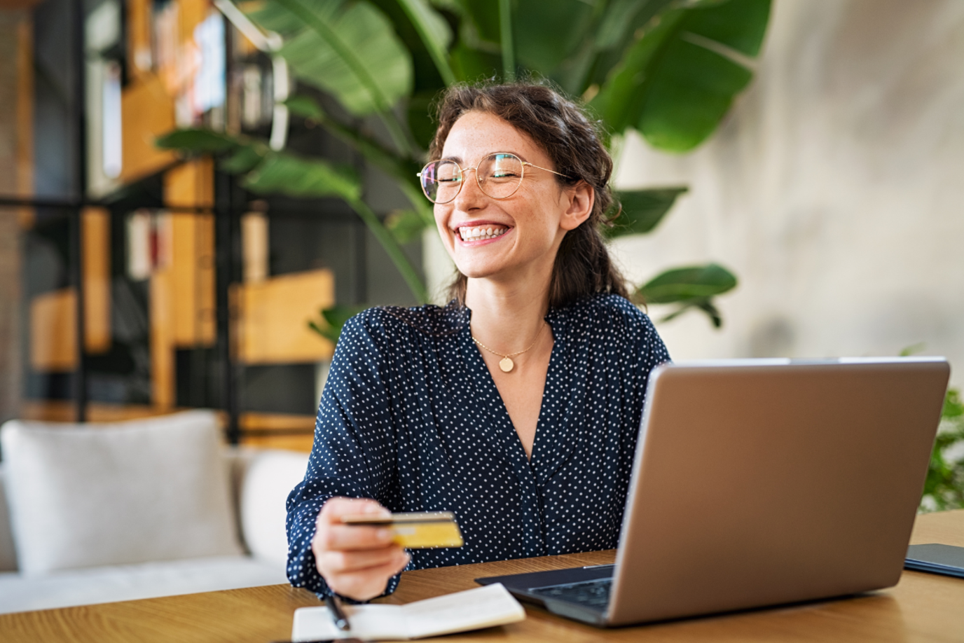 Junge Frau am Tisch mit Laptop, die glücklich lächelnd ihre Bankkarte in der Hand hält