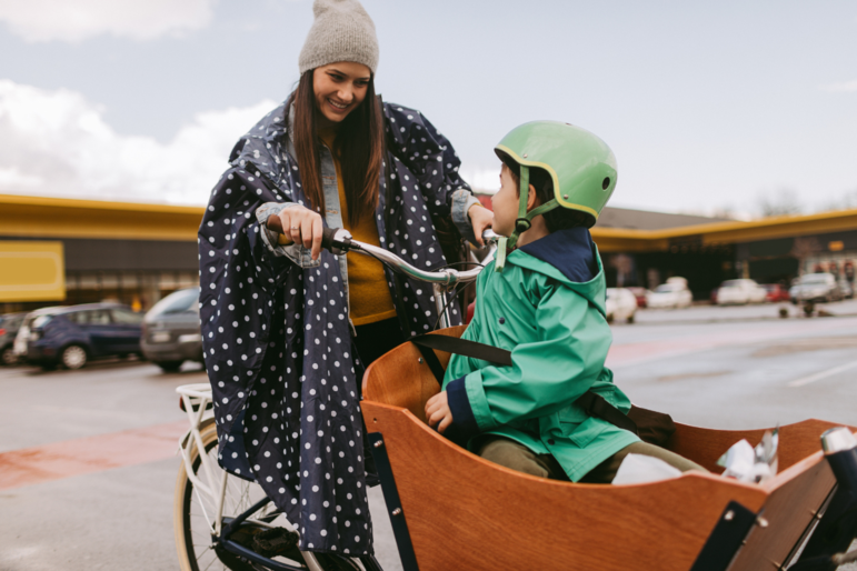 Junge Mutter mit kleinem Jungen im Lastenrad vor einem Supermarkt, die sich lächelnd anschauen