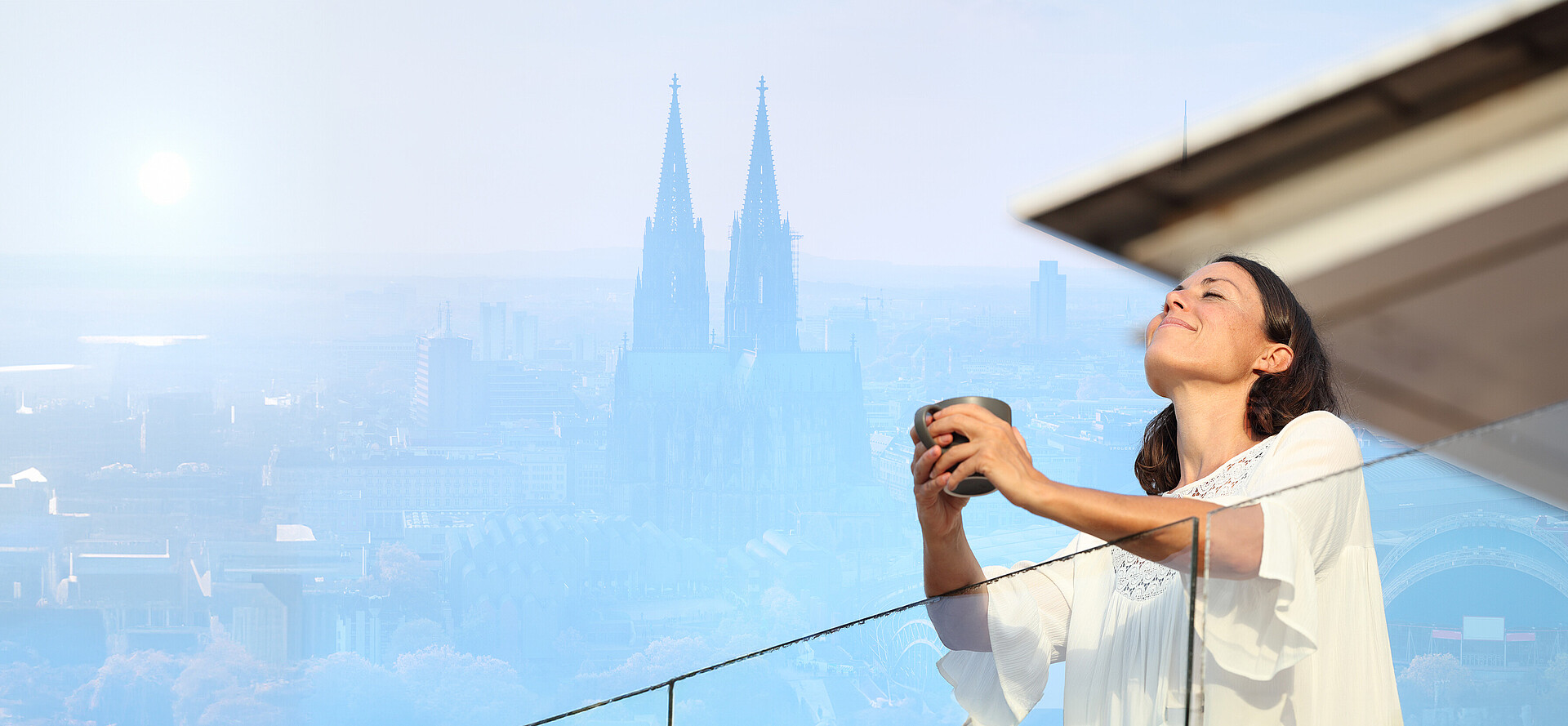 Frau steht mit einer Tasse in den Händen auf einem Balkon, mit dem Kölner Dom im Hintergrund