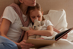 Eine Mutter hält ihre Tochter vor dem Einschlafen im Arm und liest ihr vor. 