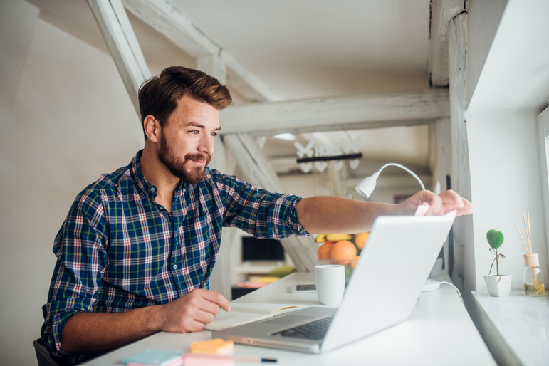 Junger Mann sitzt im Arbeitszimmer und klappt seinen Laptop auf