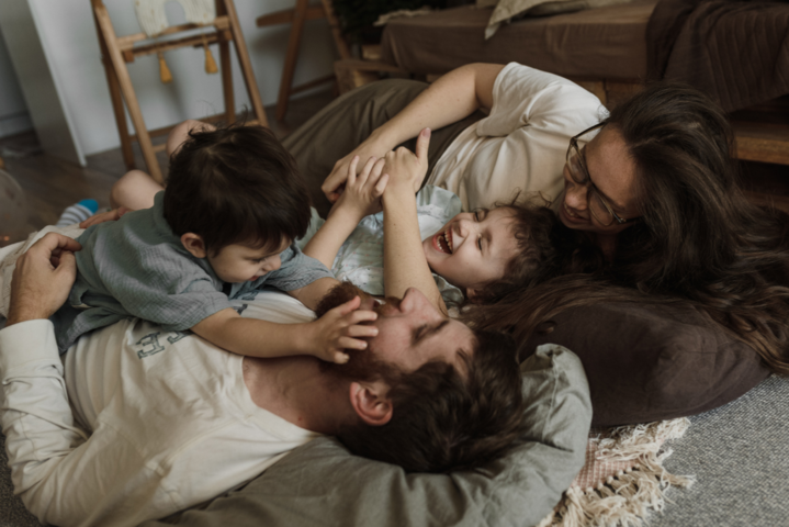 Junge Familie, bestehend aus einem Mann, einer Frau und zwei kleinen Jungen, liegt lachend und spielend auf Bodenkissen im Wohnzimmer