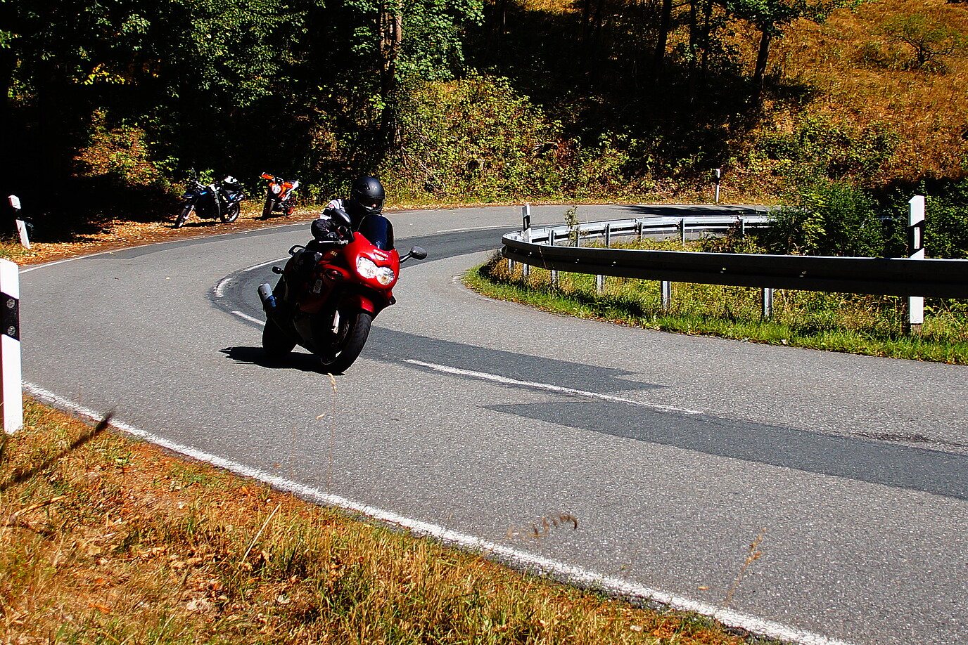 Eine motorradfahrende Person fährt eine kurvige Strecke im Harz entlang, im Hintergrund stehen zwei Motorräder