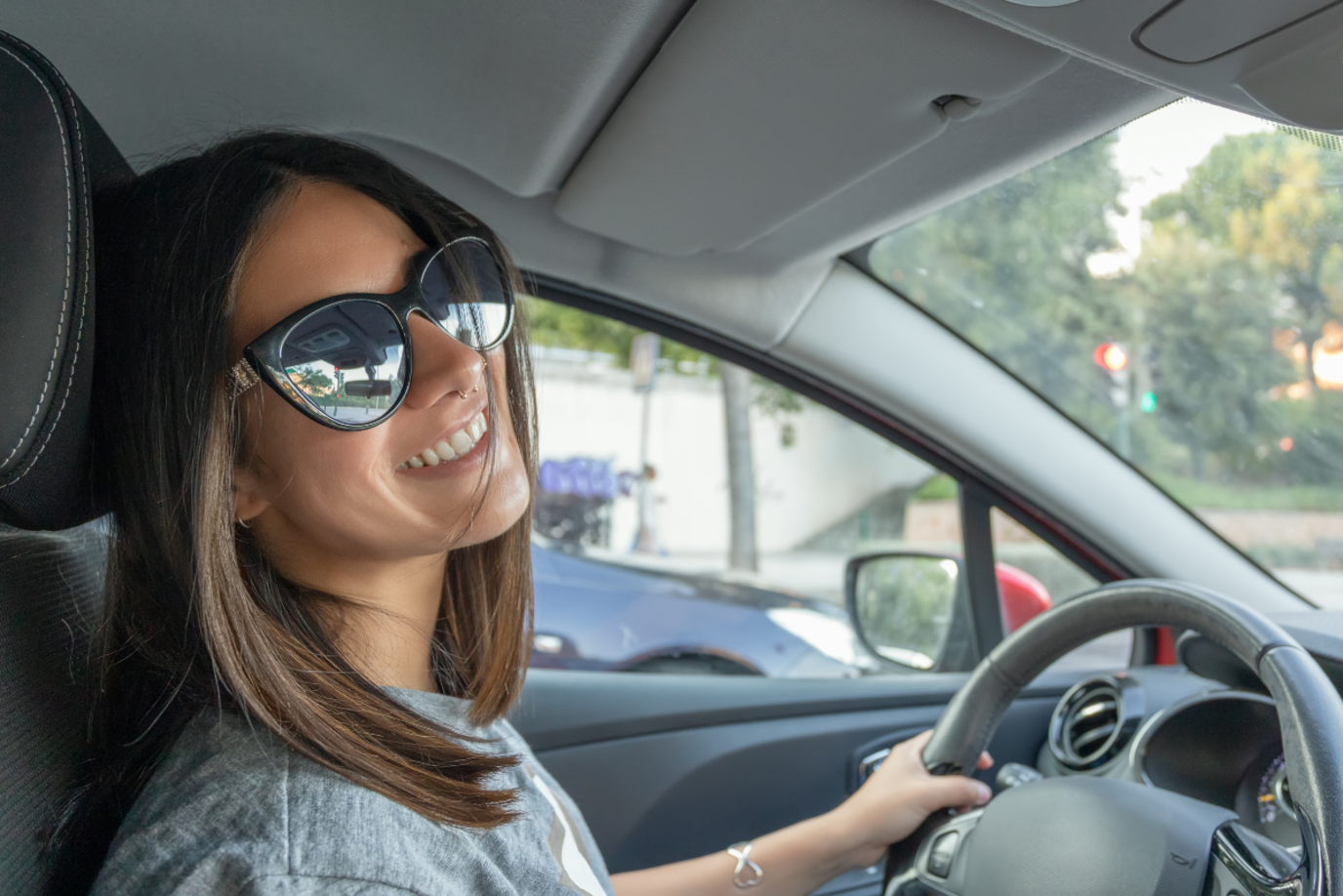 Junge Autofahrerin mit Nasenpiercing und Sonnenbrille lächelt in Richtung Beifahrer die Kamera