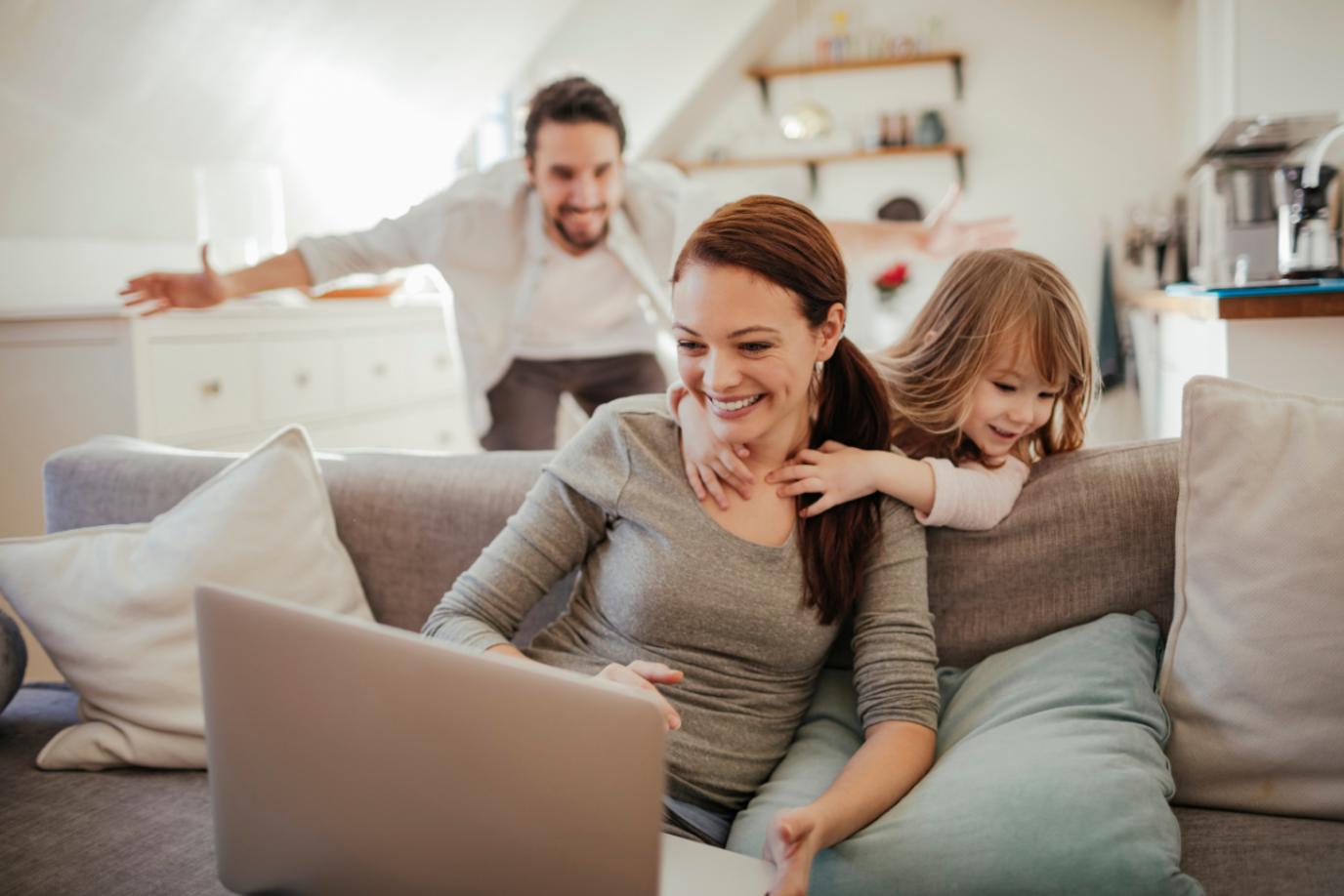 Eine junge Frau sitzt lachend auf dem Sofa mit einem Laptop, hinter ihr sind spielend ihre kleine Tochter und ihr Mann