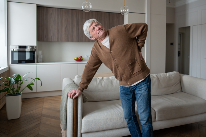 Rentner mit Rückenschmerzen im Wohnzimmer, der sich gequält an den Rücken fasst