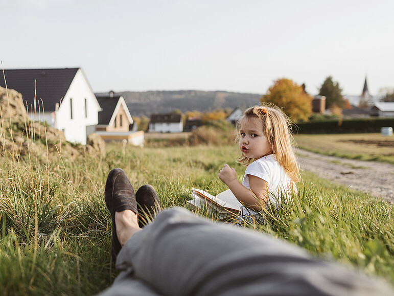 Portrait eines kleinen Mädchens, das mit einem Buch auf einer Wiese sitzt und ihre Mutter anschaut.