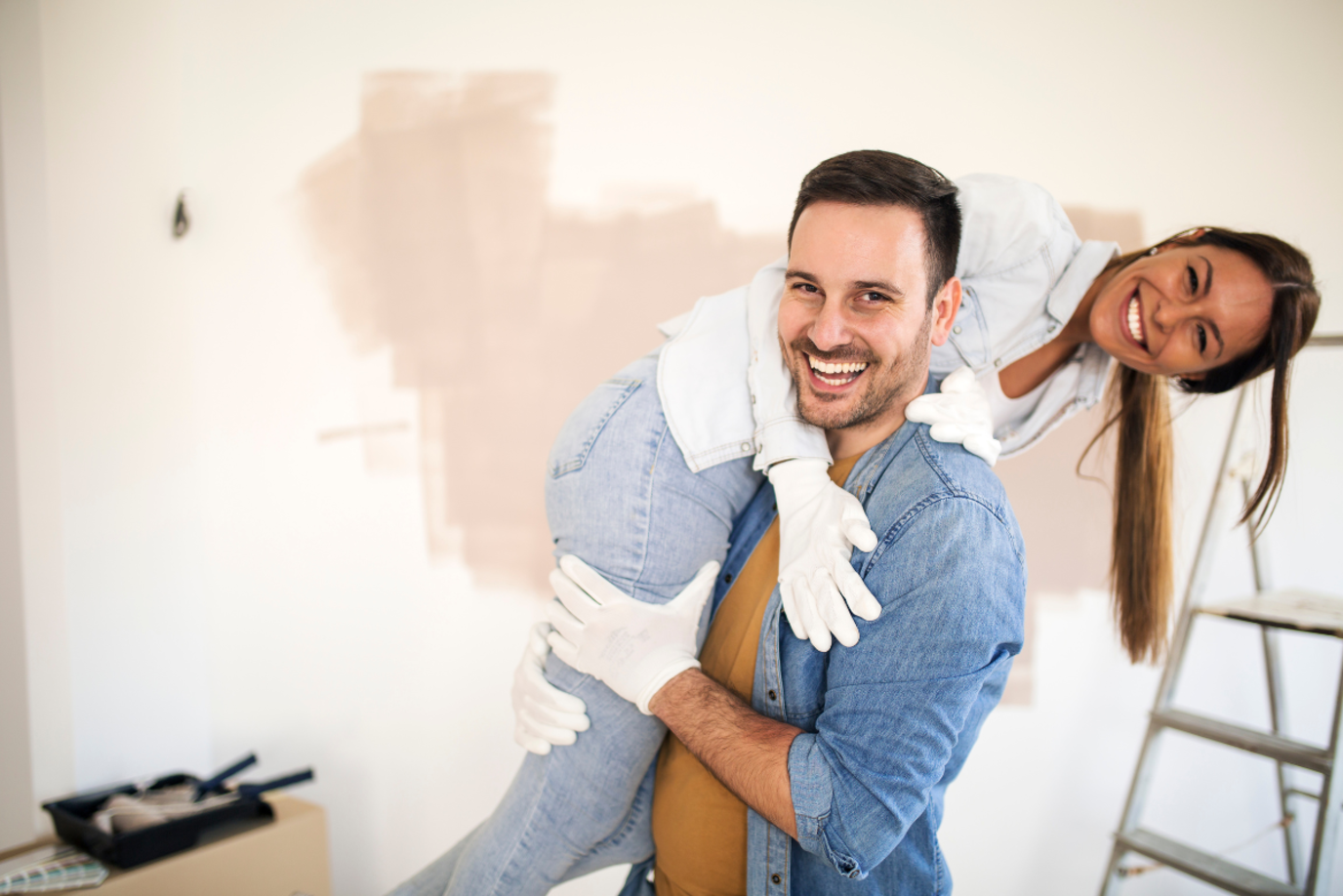 Ein glückliches Pärchehen bestehend aus einem Mann und einer Frau beim Renovieren. Der Mann hebt die Frau aus Spaß hoch, beide Lachen