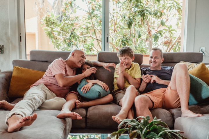 Zwei herzhaft lachende Männer mit ihrem lachenden 12-jährigen Sohn auf dem Familiensofa im Wohnzimmer