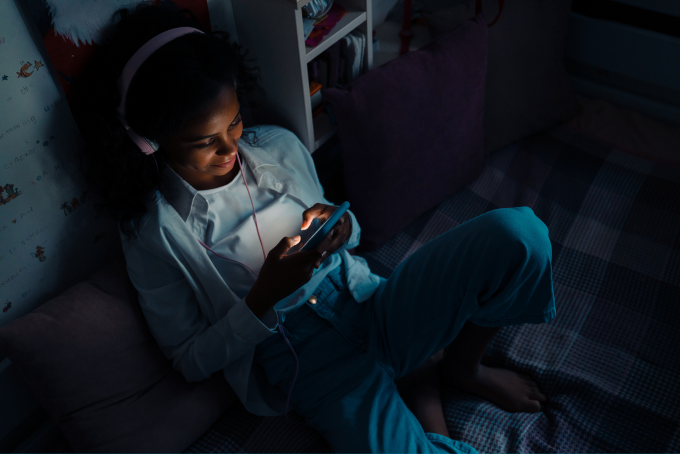 Junges Mädchen sitzt mit Kopfhörern im dunklen Kinderzimmer und schaut auf ihr Handy