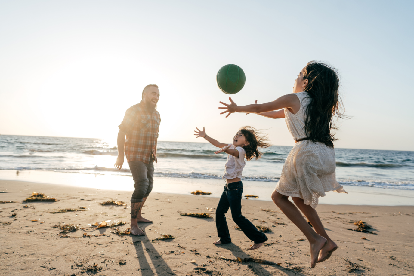Ein Vater spielt bei Sonnenuntergang am Strand mit seinen beiden Töchtern im Alter von etwa 10 Jahren Ball; sie werfen und fangen den Ball mit den Händen