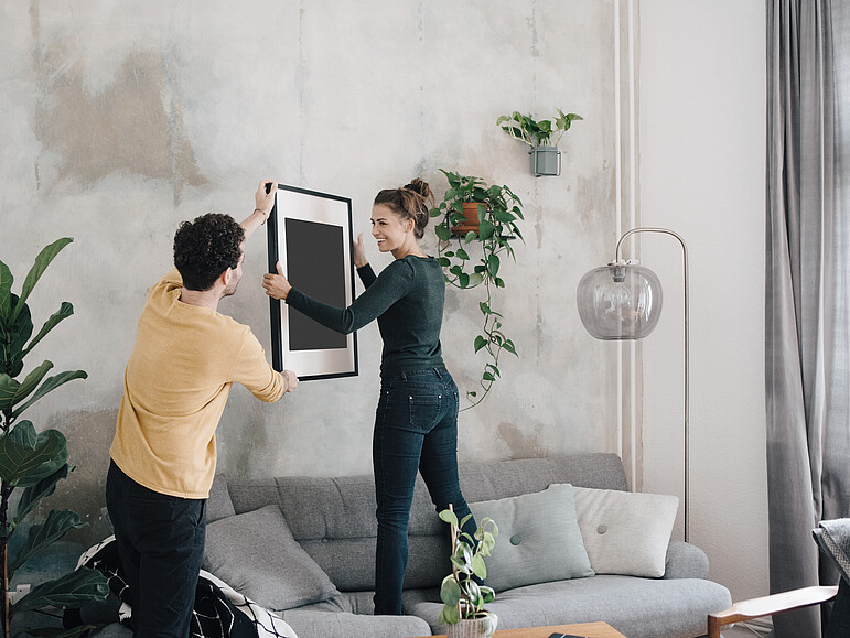 Erwachsener Mann reicht seiner Freundin einen Bilderrahmen an die Wand vom neuen Haus  