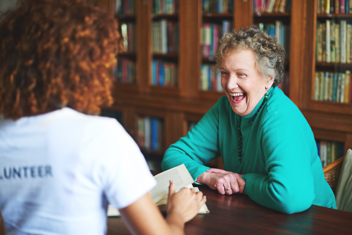 Junge Frau sitzt mit einer älteren Dame am Tisch; beide lachen herzhaft
