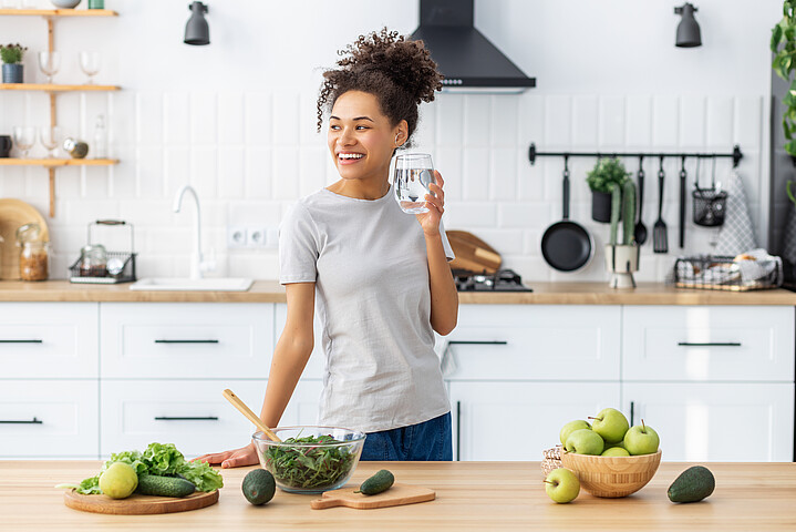 Eine junge Frau steht in ihrer Küche und hält lächelnd ein Glas Wasser.