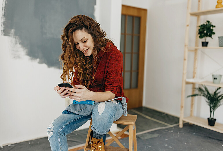 Frau macht auf einem Hocker sitzend Pause vom Streichen eines Raumes und hält ihr Handy lächelnd in der Hand