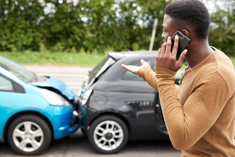 Junger BIPoC Mann am Telefon meldet Schaden nach einem Auto-Auffahrunfall