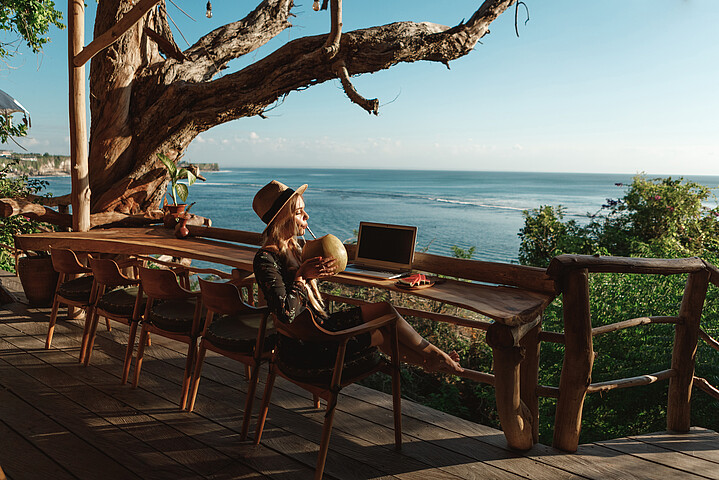 Eine junge Frau arbeitet im Ausland am Laptop und sitzt entspannt auf einer Terrasse.