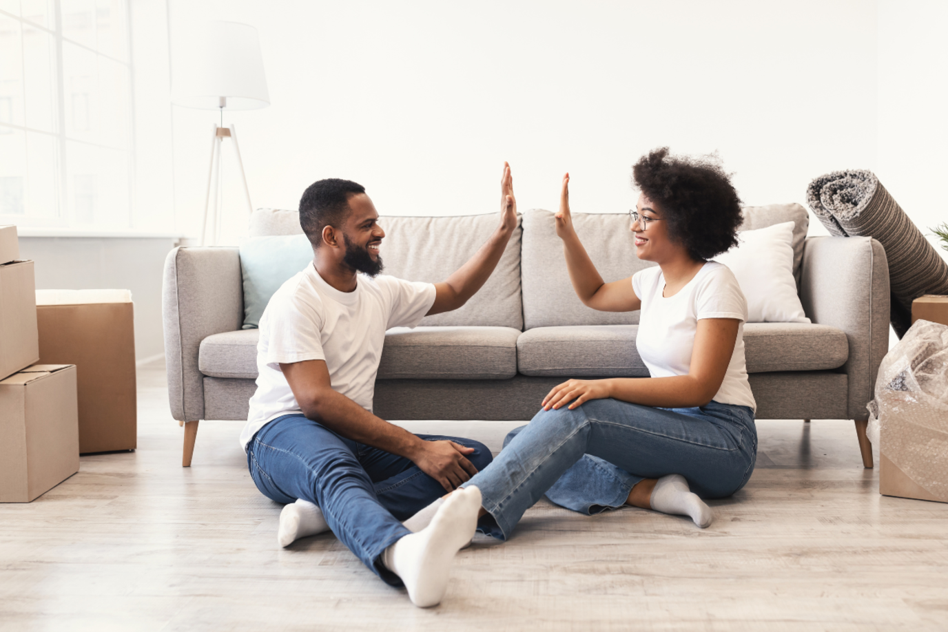 Ein Mann und eine Frau sitzen zufrieden vor einem Sofa auf dem Boden und klatschen sich ab. Links und rechts stehen Umzugskartons