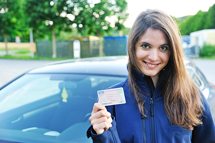 Junge Fahranfängerin hält lachend ihren Führerschein in der Hand. 