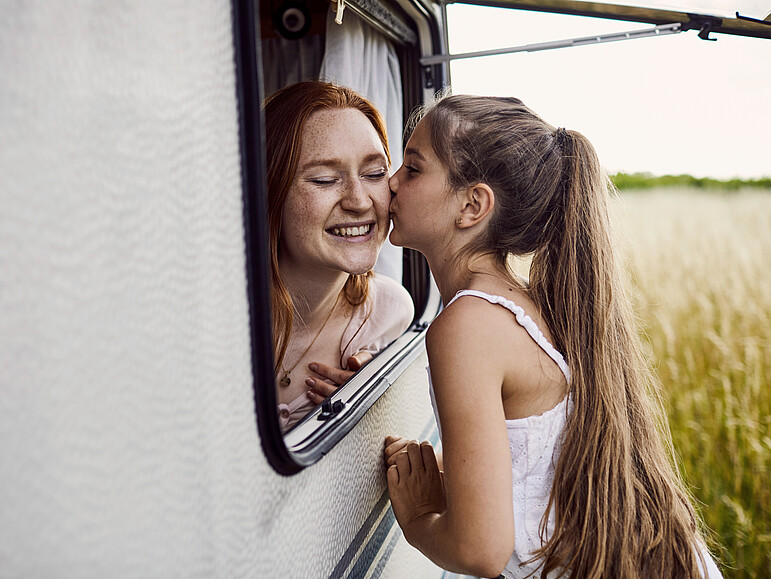 Ein Mädchen gibt ihrer älteren Schwester auf die Wange einen Kuss durch das geöffnete Fenster eines Wohnwagens.