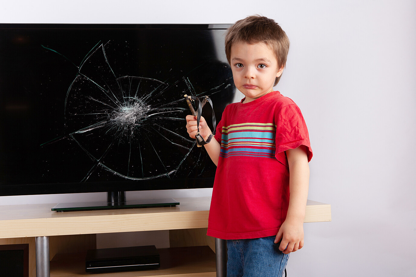 Ein Kind steht mit einer Zwille vor einem zerstörten Fernsehgerät