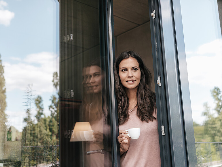 Lächelnde Frau schaut aus einer halb geöffneten Balkontür und hält dabei eine Tasse in der Hand.