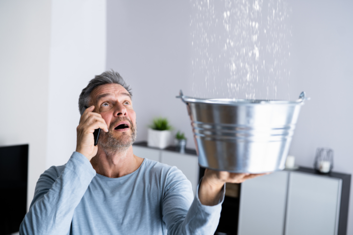 Erschrockener Mann am Handy, der von der Decke tropfendes Wasser mit einem Eimer auffängt