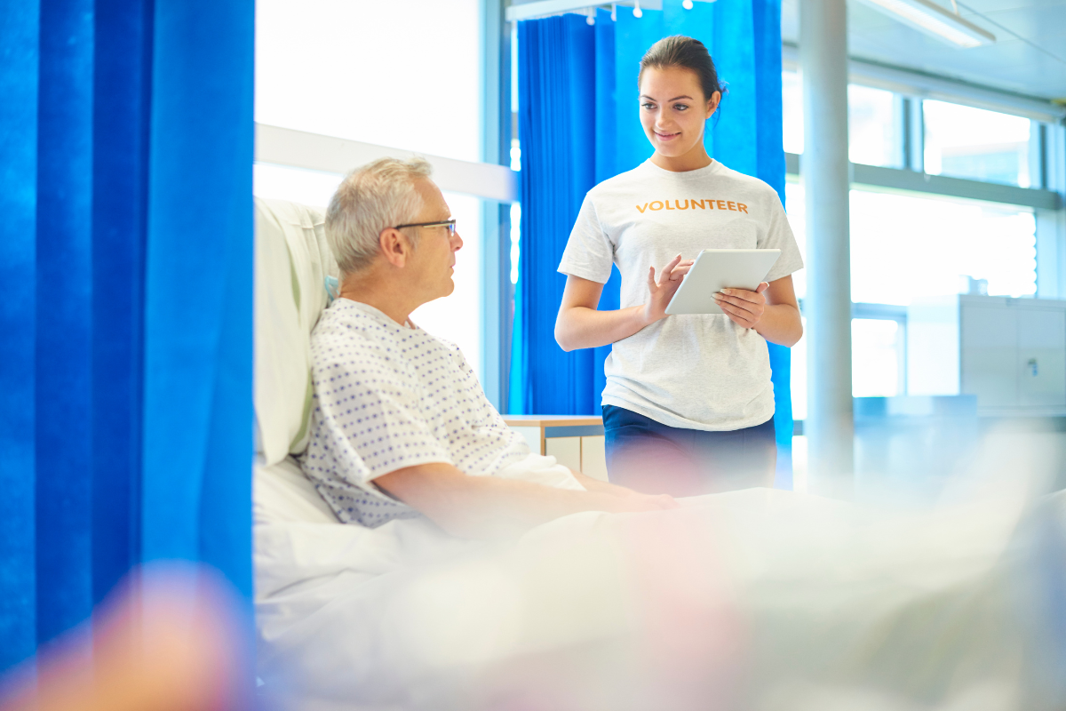 Eine junge, ehrenamtliche Helferin unterhält sich mit einem älteren Herrn am Krankenhausbett Eine junge, ehrenamtliche Helferin steht vor dem Krankenbett eines älteren Herren und unterhält sich mit ihm