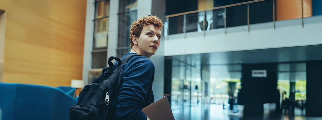 Ein Junge in einem Bürogebäude aus Glas schaut über seine rechte Schulter nach hinten und hält einen Laptop in der Hand.