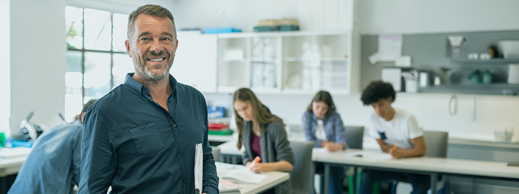 Ein Lehrer steht im Klassenzimmer, im Hintergrund arbeiten Schüler an ihren Aufgaben.