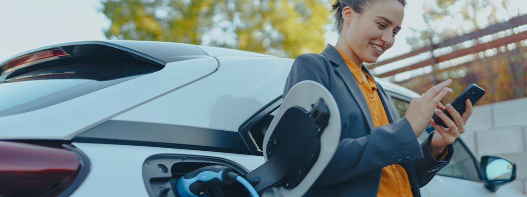 Eine Frau schaut glücklich auf ihr Handy, während ihr E-Auto aufgeladen wird.