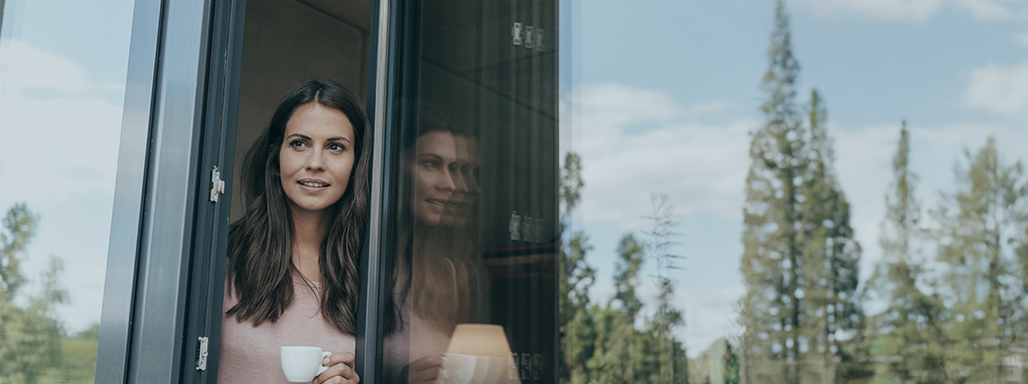 Eine junge Frau trinkt Kaffee und schaut aus einem offenen Fenster nach draußen.