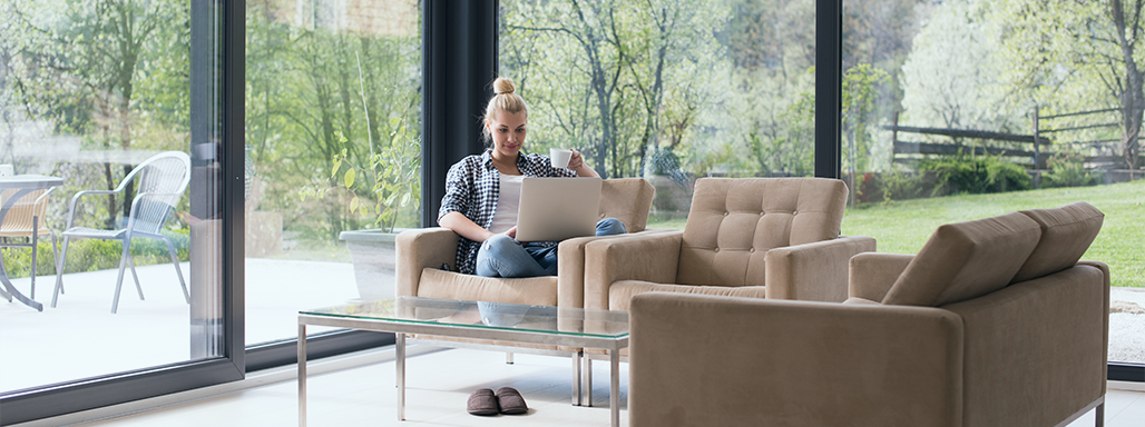 Ein Frau sitzt mit Laptop und Kaffee auf einem Sessel umgeben von großen Fenstern.