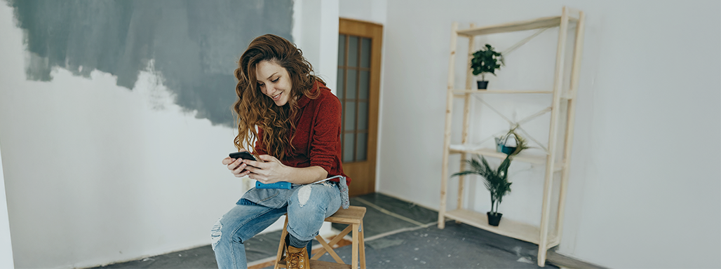 Frau macht auf einem Hocker sitzend Pause vom Streichen eines Raumes und hält ihr Handy lächelnd in der Hand.