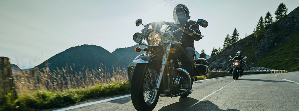 Zwei Mottoradfahrer fahren auf einer Straße mit Bergen im Hintergrund.