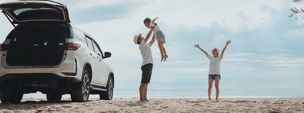 Familie am Strand neben weißem Auto, der Mann hebt das Kind hoch und die Frau jubelt.
