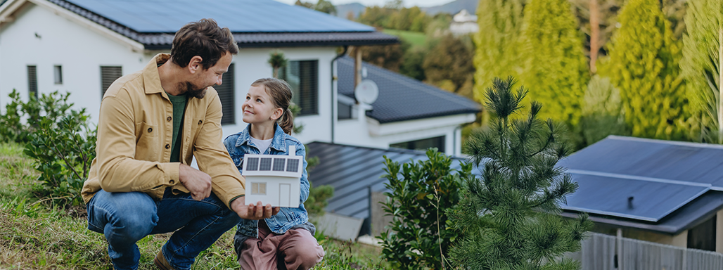 Vater und Tochter halten ein gebasteltes Haus mit Photovoltaikanlage in der Hand. Im Hintergrund sind Häuser mit Photovoltaikanlagen. 