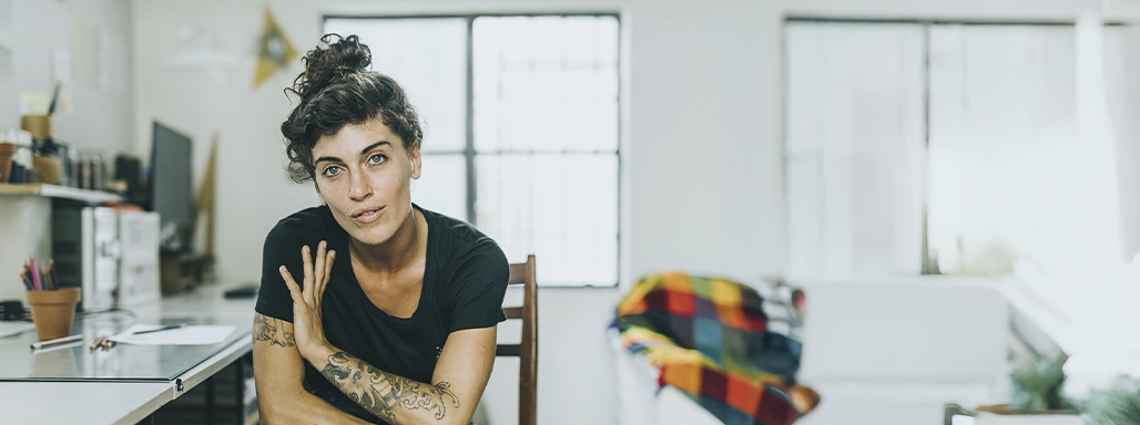 Tattoovierte Frau sitzt in der Küche auf einem Stuhl und blickt in die Kamera.