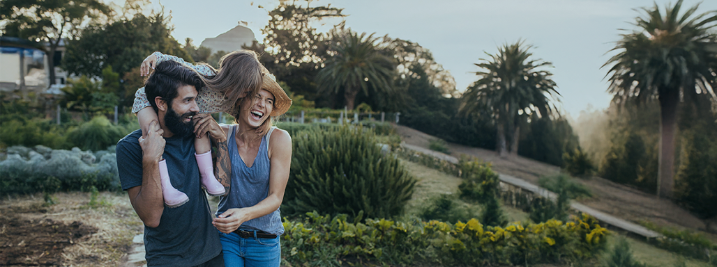 Glückliche Familie im Garten bestehend aus einem Mann, einer Frau und einem Kleinkind auf den Schultern des Vaters.