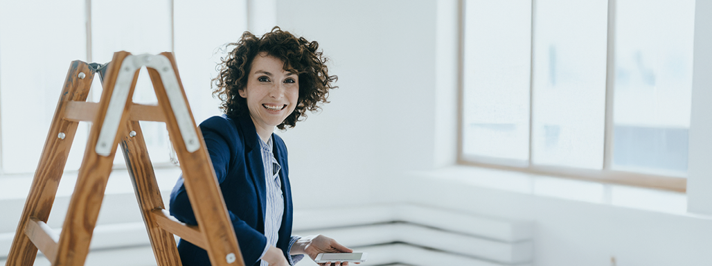 Frau sitzt im Anzug mit einem Handy auf einer Leiter in einem Wohnraum und lächelt in die Kamera.