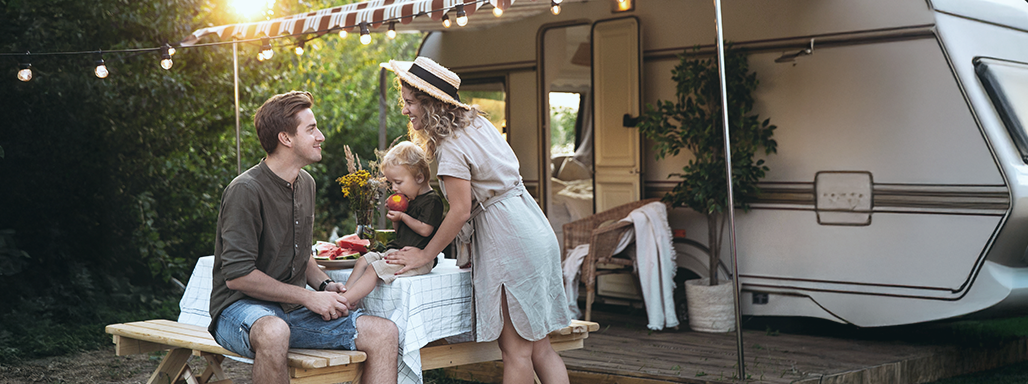 Eine junge Familie mit einem Kleinkind sitzt an einem Picknicktisch. Im Hintergrund ist ein Wohnwagen.
