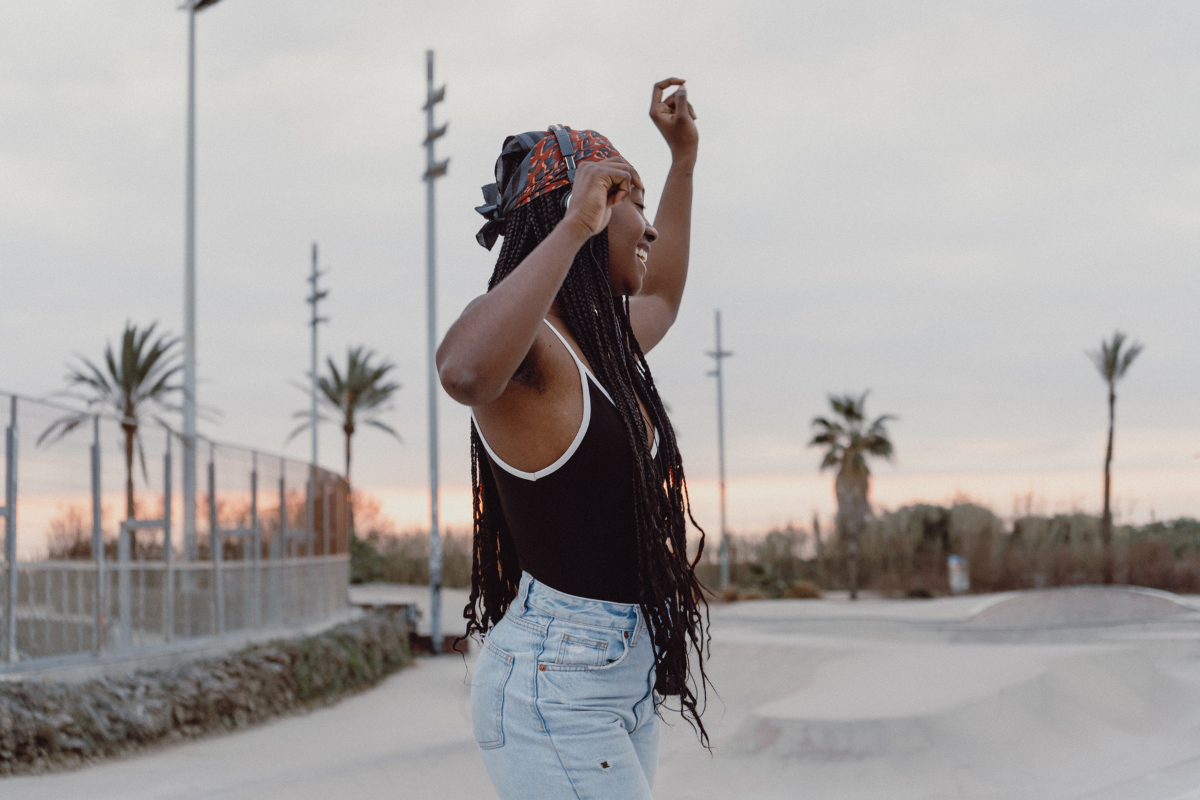 Junge BIPoC Frau tanzt draußen während einem Sonnenuntergang zur Musik
