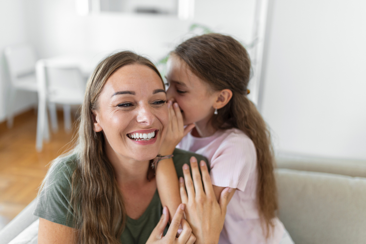 Junges Mädchen flüstert ihrer Mutter etwas ins Ohr; beide lächeln